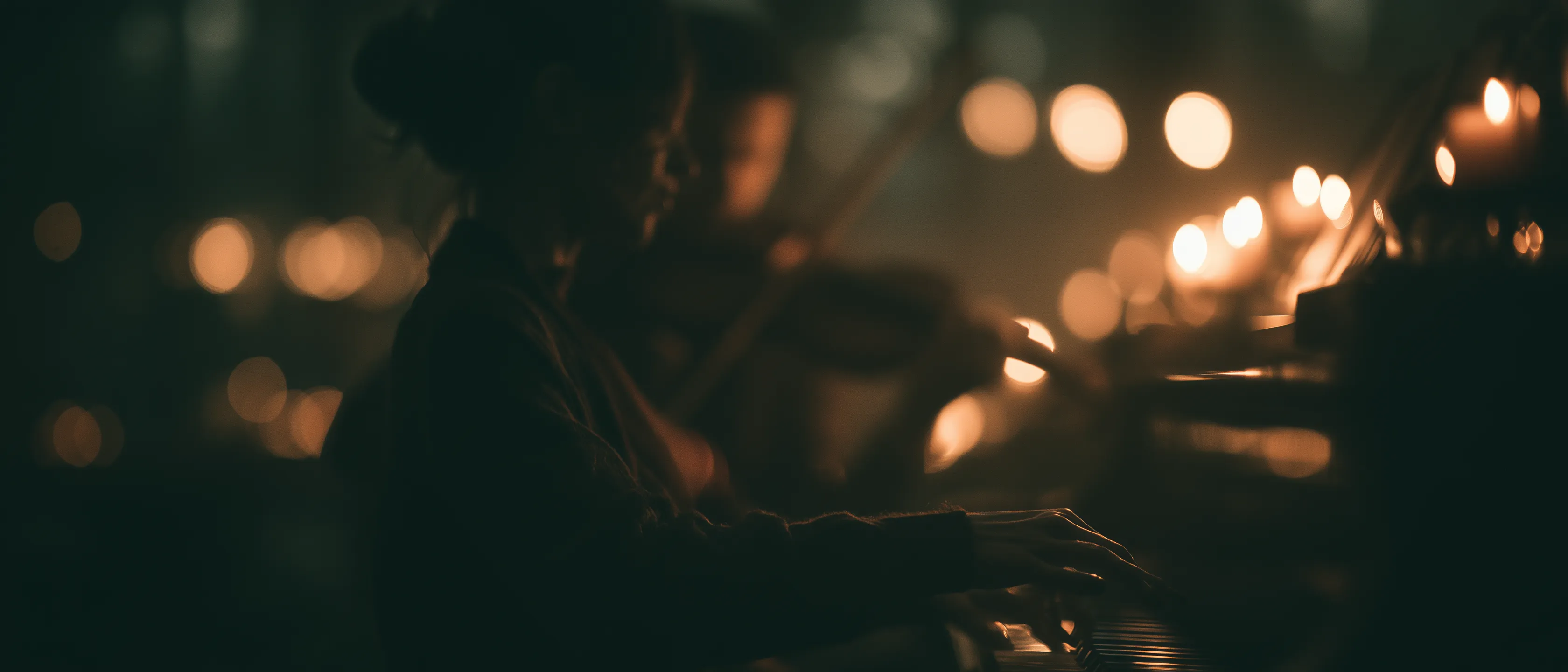 A pianist and violinist play together in near-darkness, lit only by warm rows of candlelight glowing behind them.